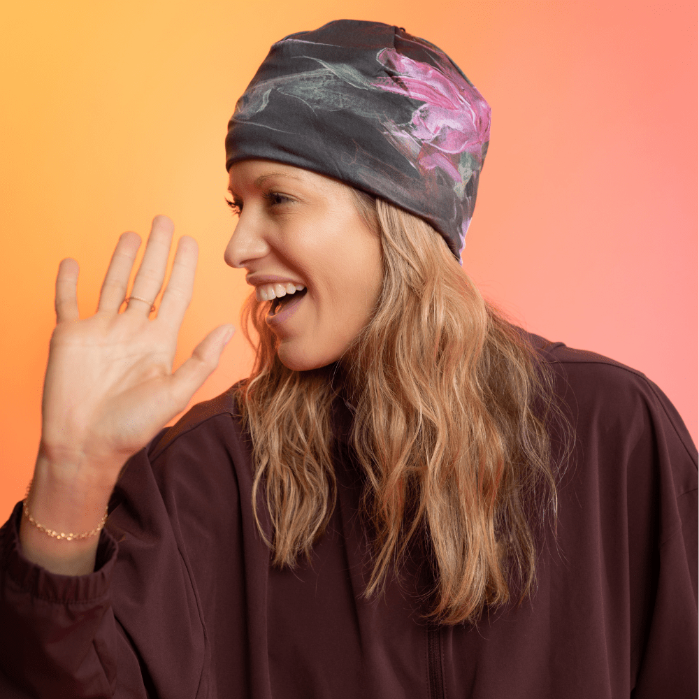 Une femme portant une tuque noire avec un imprimé floral abstrait rose et vert sourit et fait signe.