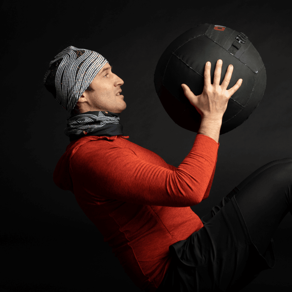 Un homme en sweat à capuche rouge portant une tuque et un cache-cou Dark Steel, tenant un gros ballon médecine.
