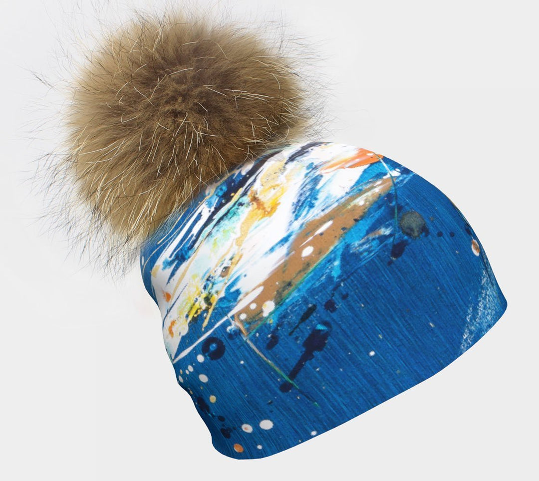Tuque bleue avec des éclaboussures de peinture abstraites colorées et un gros pompon brun et moelleux.