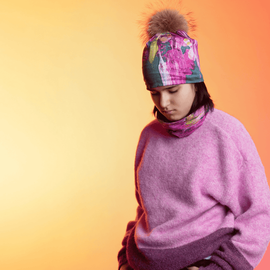 Une jeune personne portant la Tuque Fleur Glitch et une cache-cou assortie avec un chandail rose flou, regardant vers le bas sur un fond à dégradé chaud.