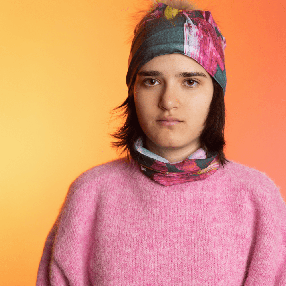 Une personne portant un chandail rose et le tube de cou Flower Glitch, avec une tuque assortie sur la tête, sur un fond dégradé.