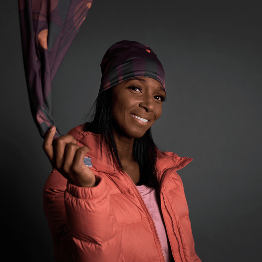 Une femme souriante en veste matelassée corail tient un bandana à motif géométrique violet, regardant vers la caméra.