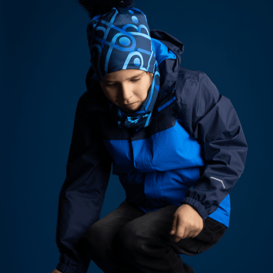 Un jeune garçon portant une veste bleue, la tuque Bleu Mécanique et un cache-cou assorti, posant dans un studio bleu foncé.