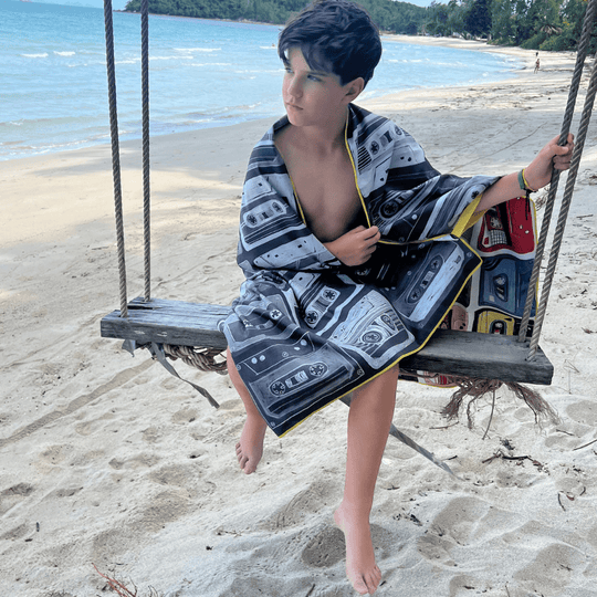 Enfant assis sur une balançoire en bois sur une plage, enveloppé dans la face noir et blanc de la serviette de plage Mixtapes.