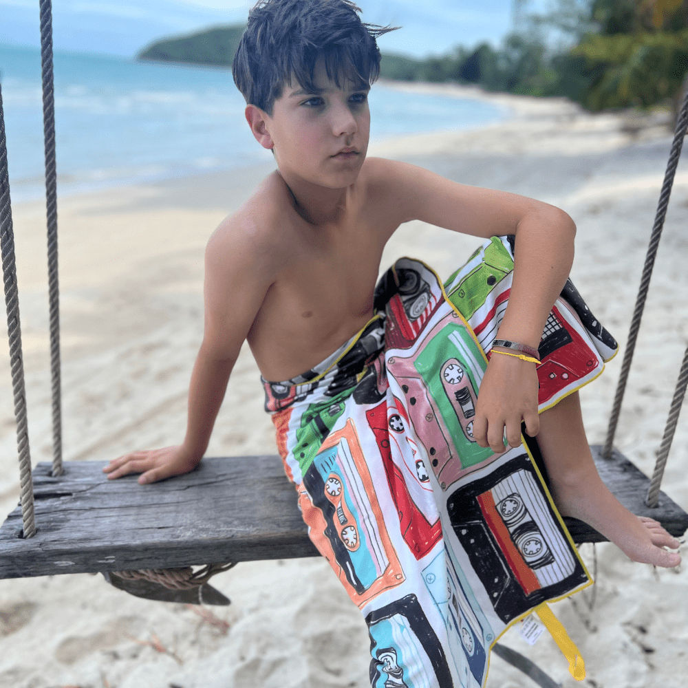 Enfant sur une balançoire en bois sur une plage, enveloppé dans la face colorée de la serviette de plage Mixtapes.