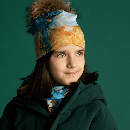 A young girl wearing a green winter coat and a matching beanie with a pompom, with the Ocean Marble tube visible around her neck.