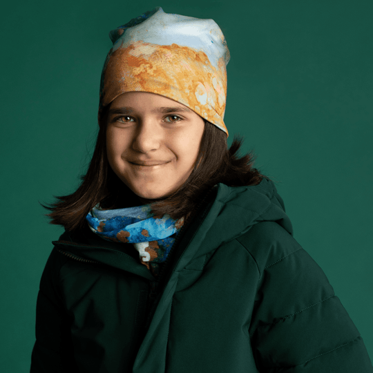 A young girl wearing a green winter coat, a blue multifunctional tube around her neck, and a matching beanie.