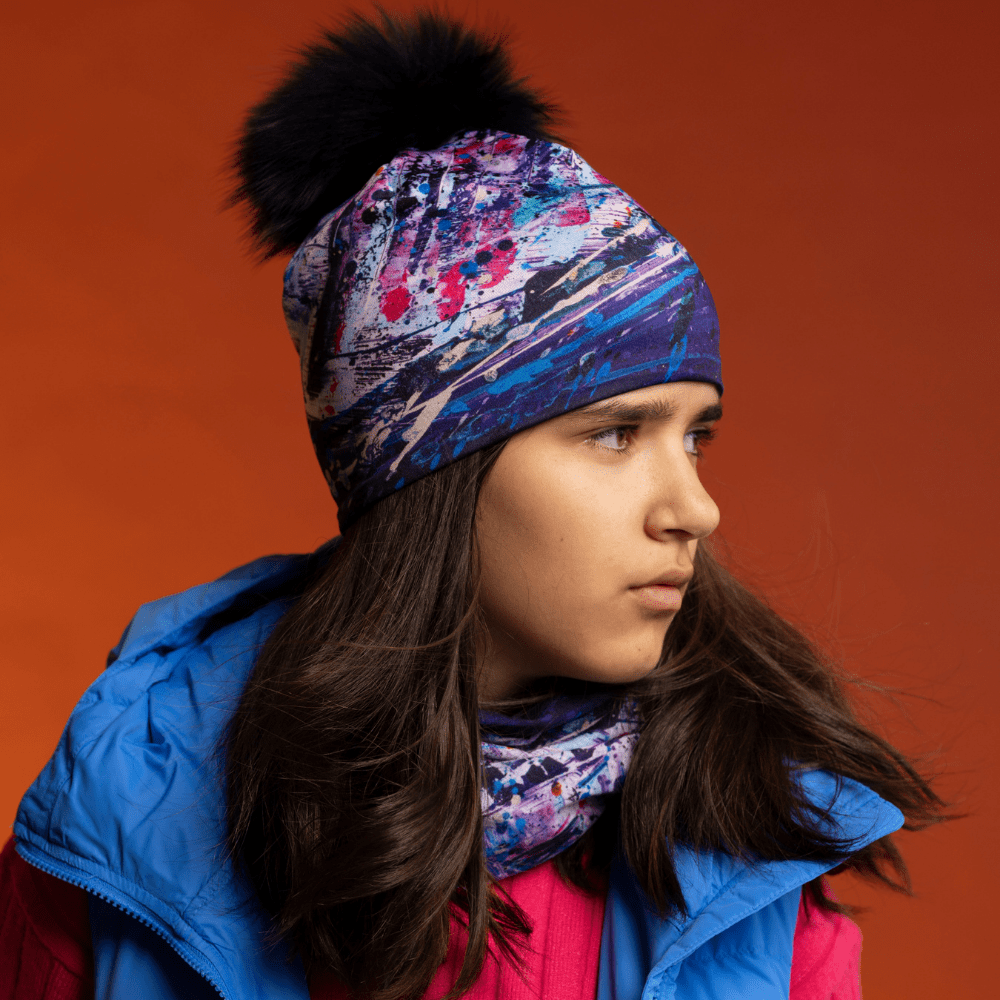 Un jeune portant une veste d'hiver bleue et un haut rose, avec la tuque et le tube cosmique de Lalita's Art Shop visibles.