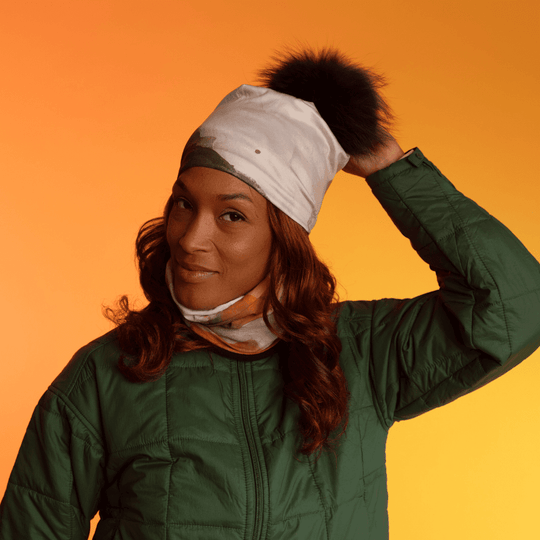 A woman wearing a green quilted jacket, adjusting a beanie adorned with a pom-pom, with the POLAR Navy Accent Multifunctional Tube visible as a neck scarf.