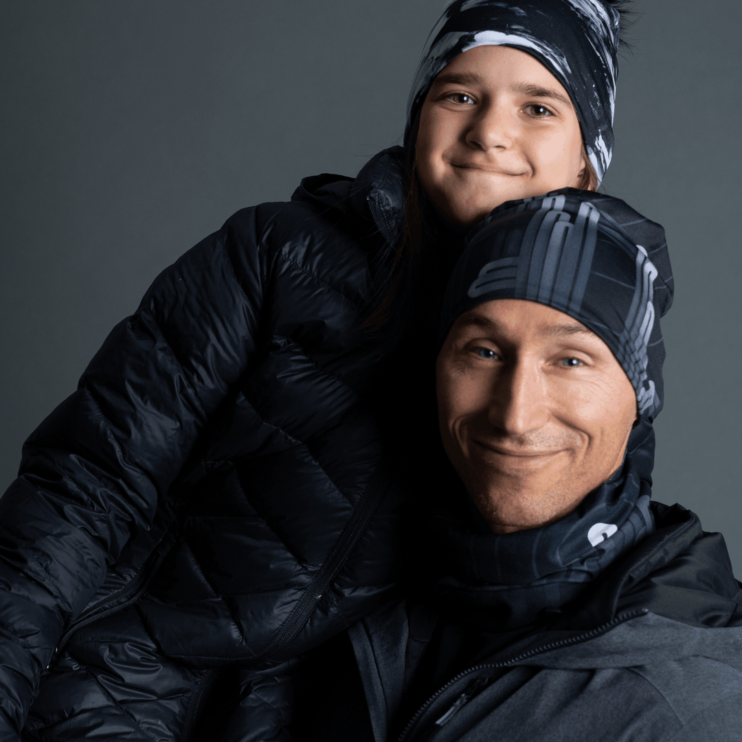 A man and a child wearing matching black beanies and neck tubes with a grey graphic design, bundled in winter jackets.