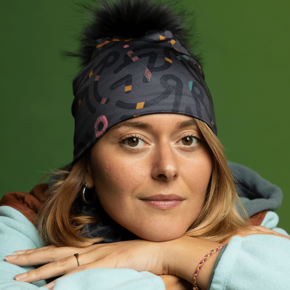 Femme portant une tuque grise avec pompon noir et cache-cou assorti, posant les mains jointes.
