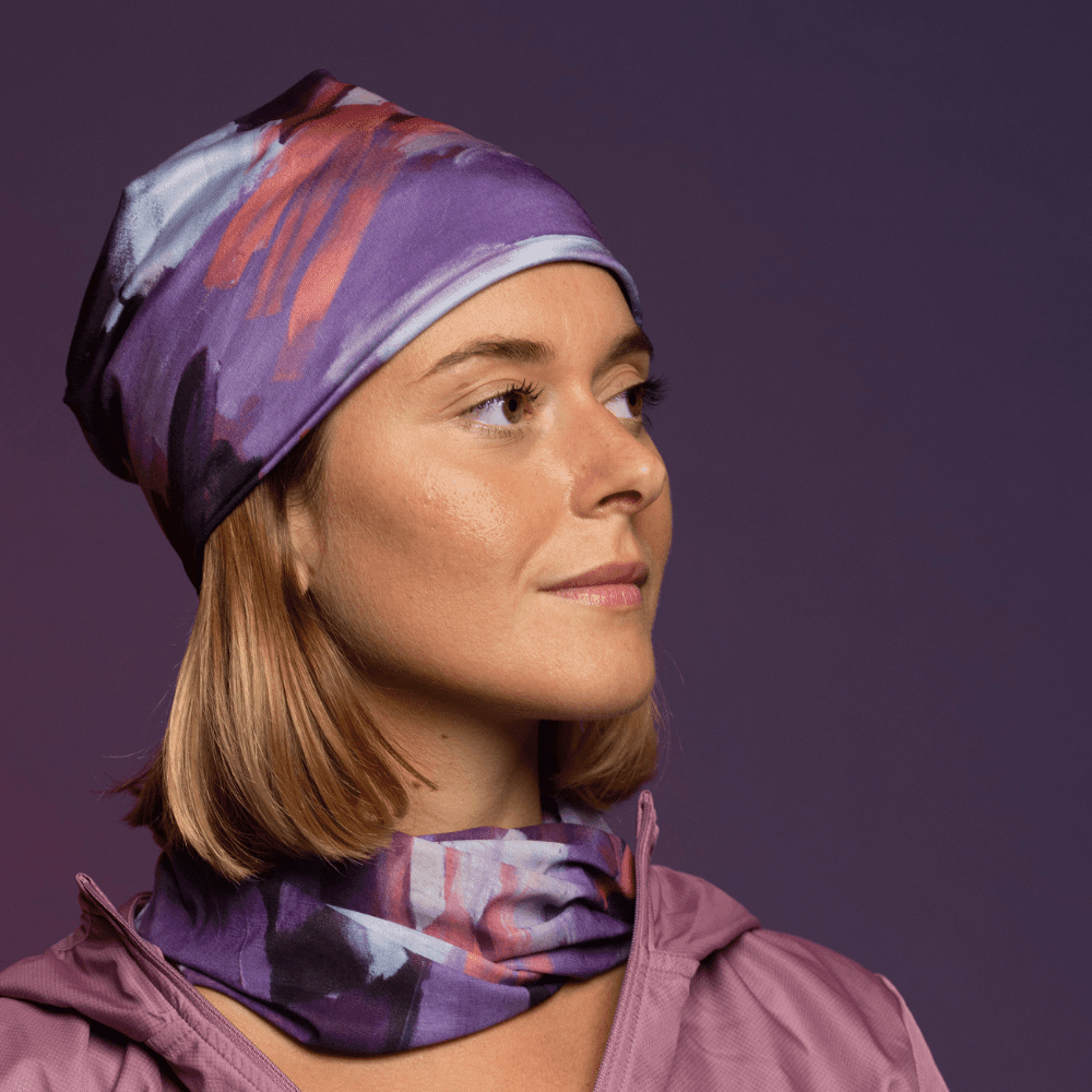 Femme portant une tuque à motif abstrait violet et un cache-cou assorti, regardant pensivement sur le côté.