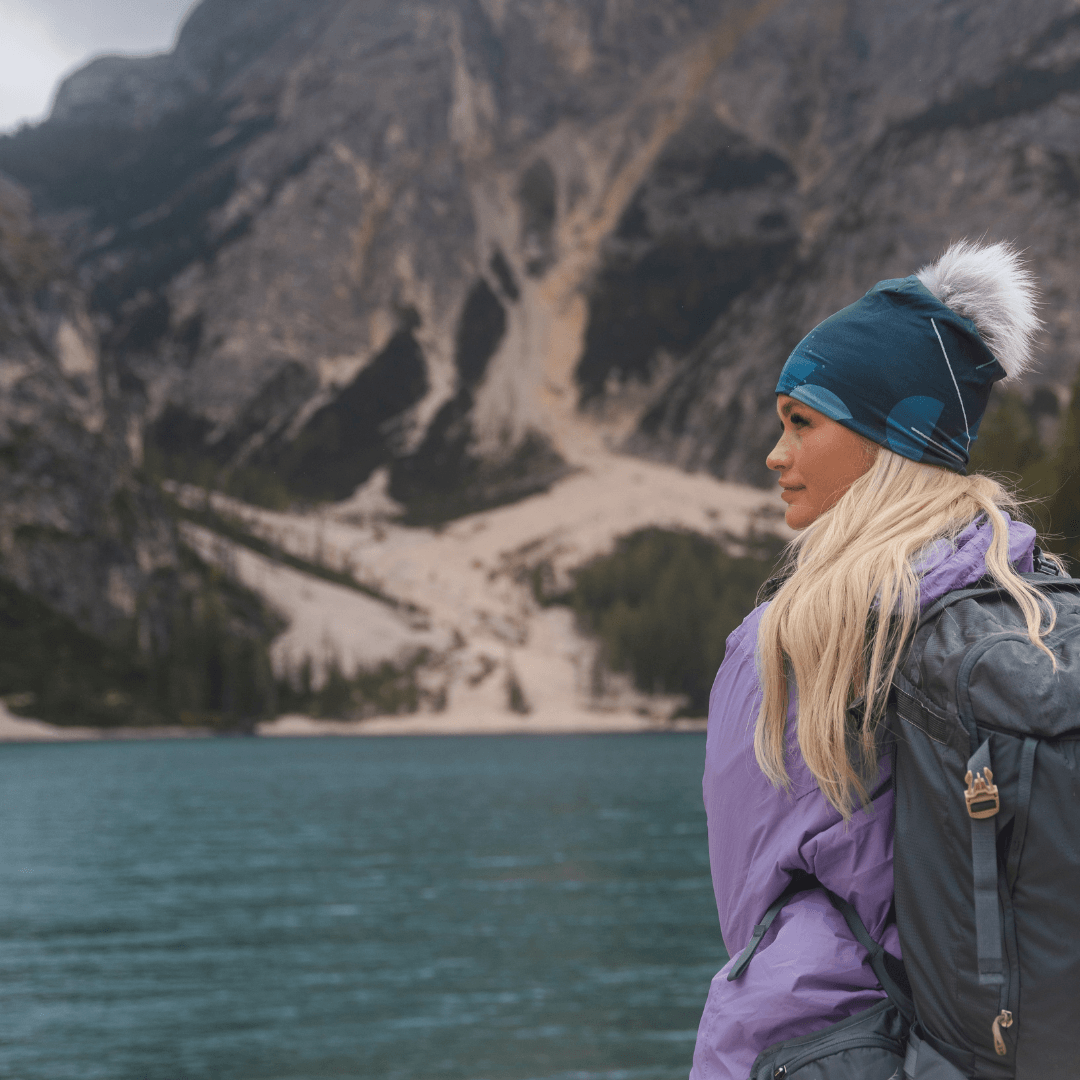 Femme portant la tuque bleu-vert avec un pompon gris et une veste violette, regardant un lac pittoresque avec des montagnes en arrière-plan.