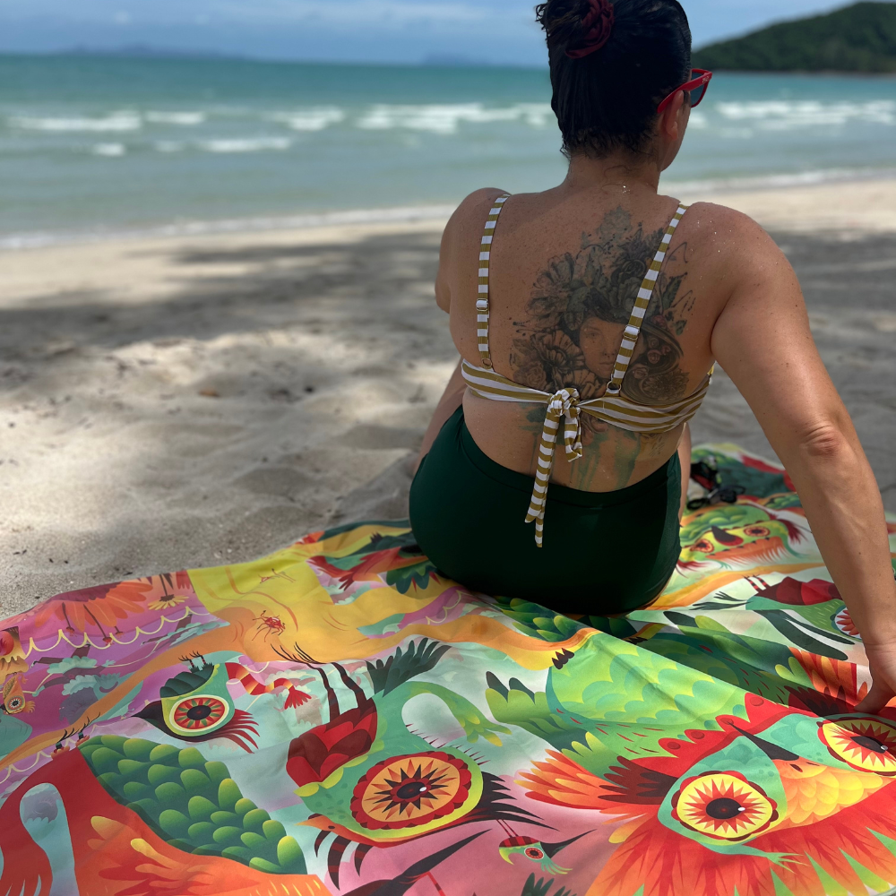 Femme tatouée assise sur une plage sablonneuse, avec le côté coloré et illustré de la serviette réversible Mur d&