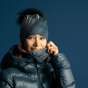 Blond woman weraing the blanck, white and blue pompom beanie hat with a dark gray jacket. 