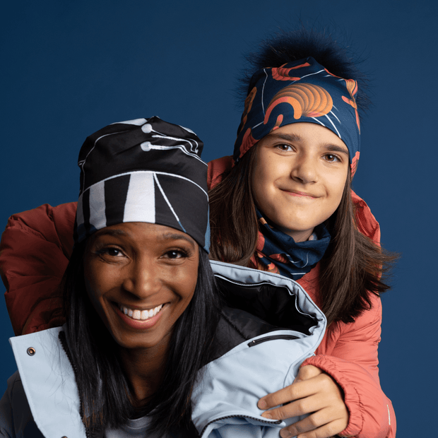 Smiling woman wearing the black and gray beanie designed by the artist Valery Goulet. This unisex beanie can be worn in any season and is perfect for the whole family. A young girl is behind the lady and smiling. Our Lalita's Art Shop hats are designed to go everywhere with you!