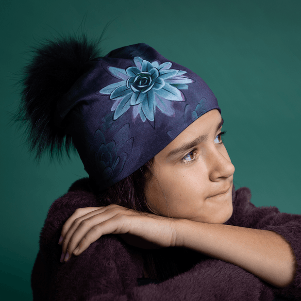 Young girl with her arms crossed and her face to the side wearing the Succulent beanie designed by Claire Anghinophi. This yoga and sports beannie hat is in purple tones with flowers designed and has a black pom-pom.