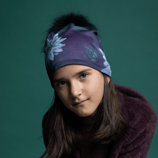 Young girl with face on, wearing the Succulent beanie designed by Claire Anghinophi. This yoga and sports beannie hat is in purple tones with flowers designed and has a black pom-pom.