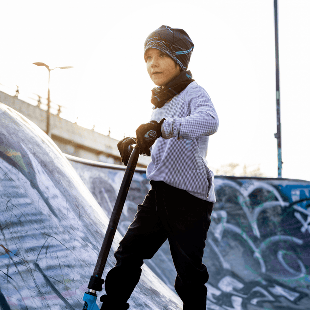Young boy in a skate park riding a scooter looking far away. The young boy is wearing a protective helmet over the Robocat cap designed by the artist André Martel